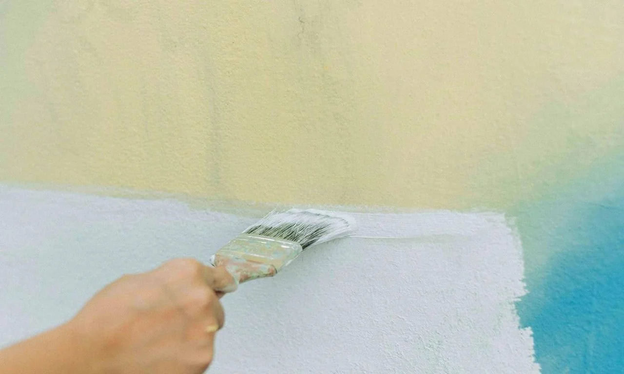 A hand using a paintbrush to apply light-colored paint to a wall.